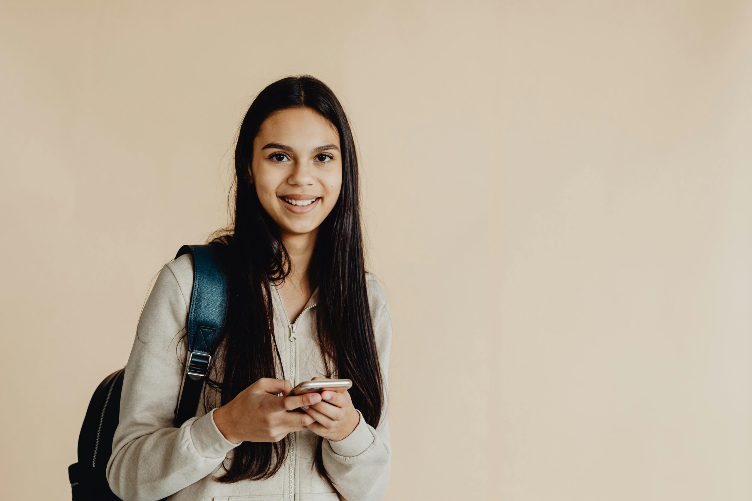 Teen girl with backpack using a smartphone against a beige background, smiling.