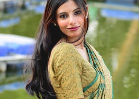 A woman in a green sari poses gracefully by a pond with boats in the background.